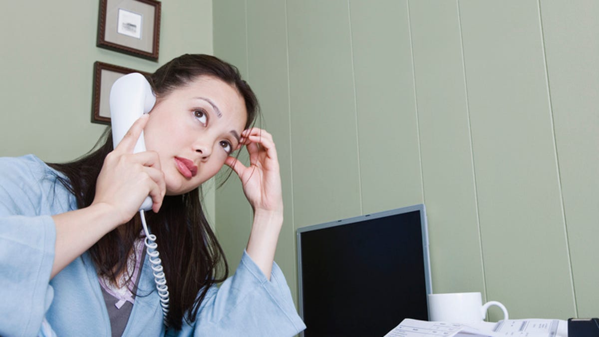 A woman looks into the distance with her hand on her head while on the phone at her desk.