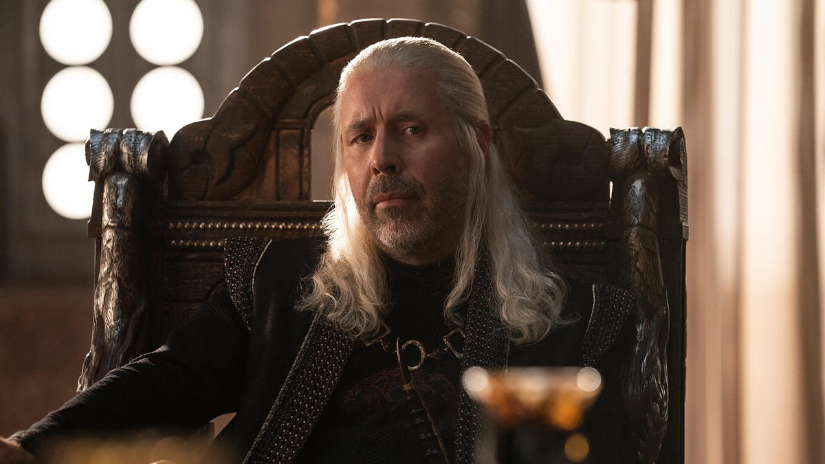 King Viserys Targaryen, seated in a heavy wooden chair, with a pensive look