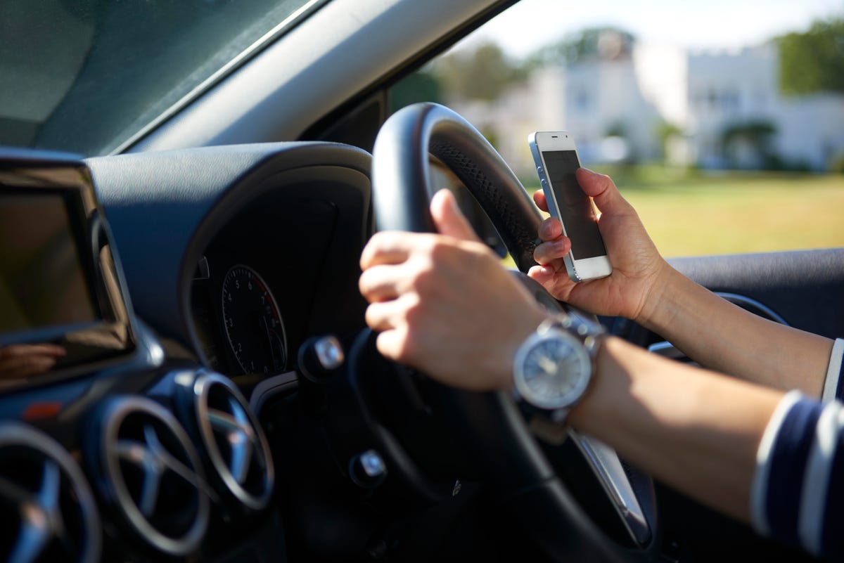 A driver with one hand on the steering wheel and the other holding a cellphone
