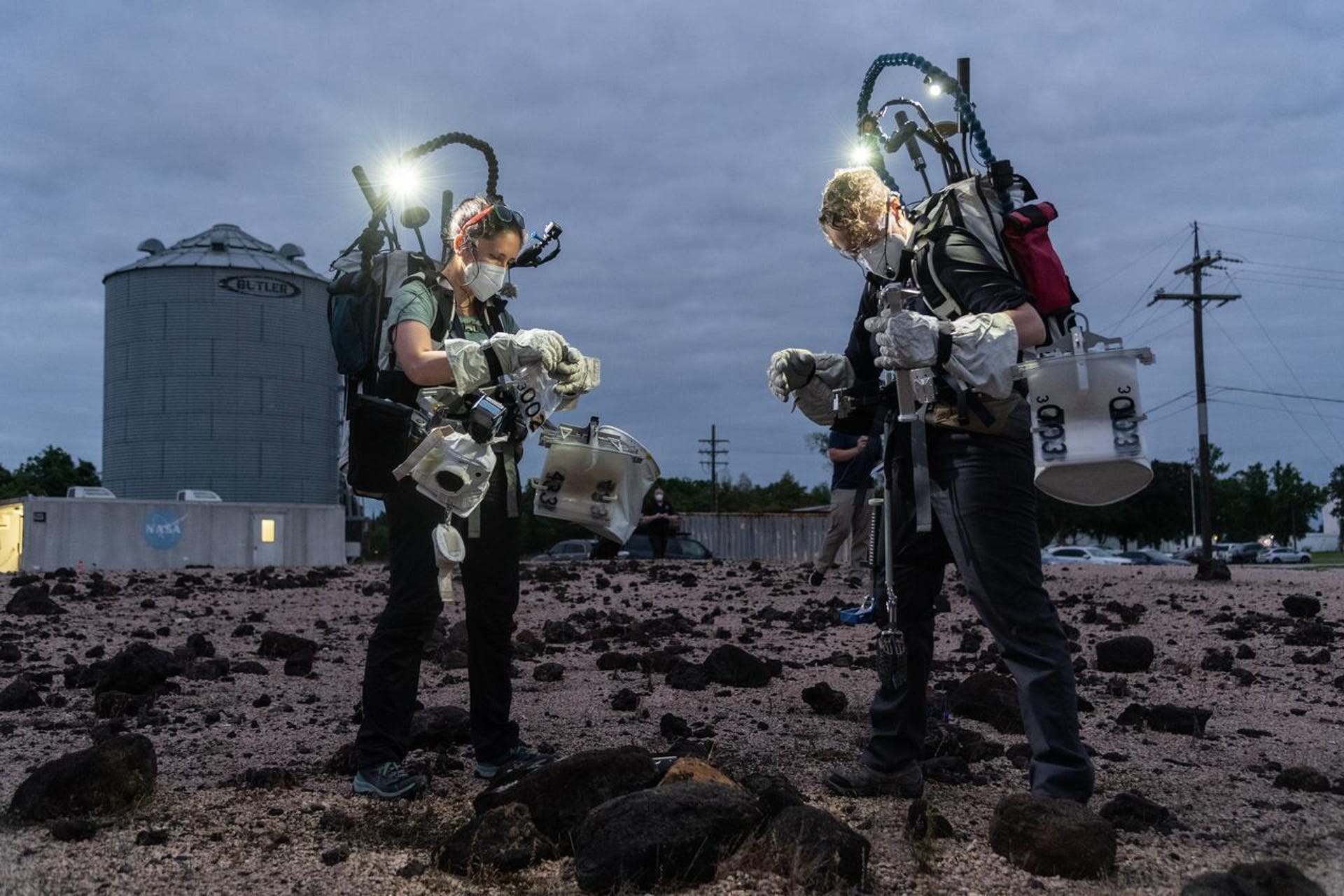 Two people wear geology testing gear in a rocky field at night with lights shining on their work.