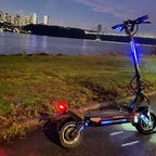 An electric scooter parked on the roadside at night with city skyline in the background.