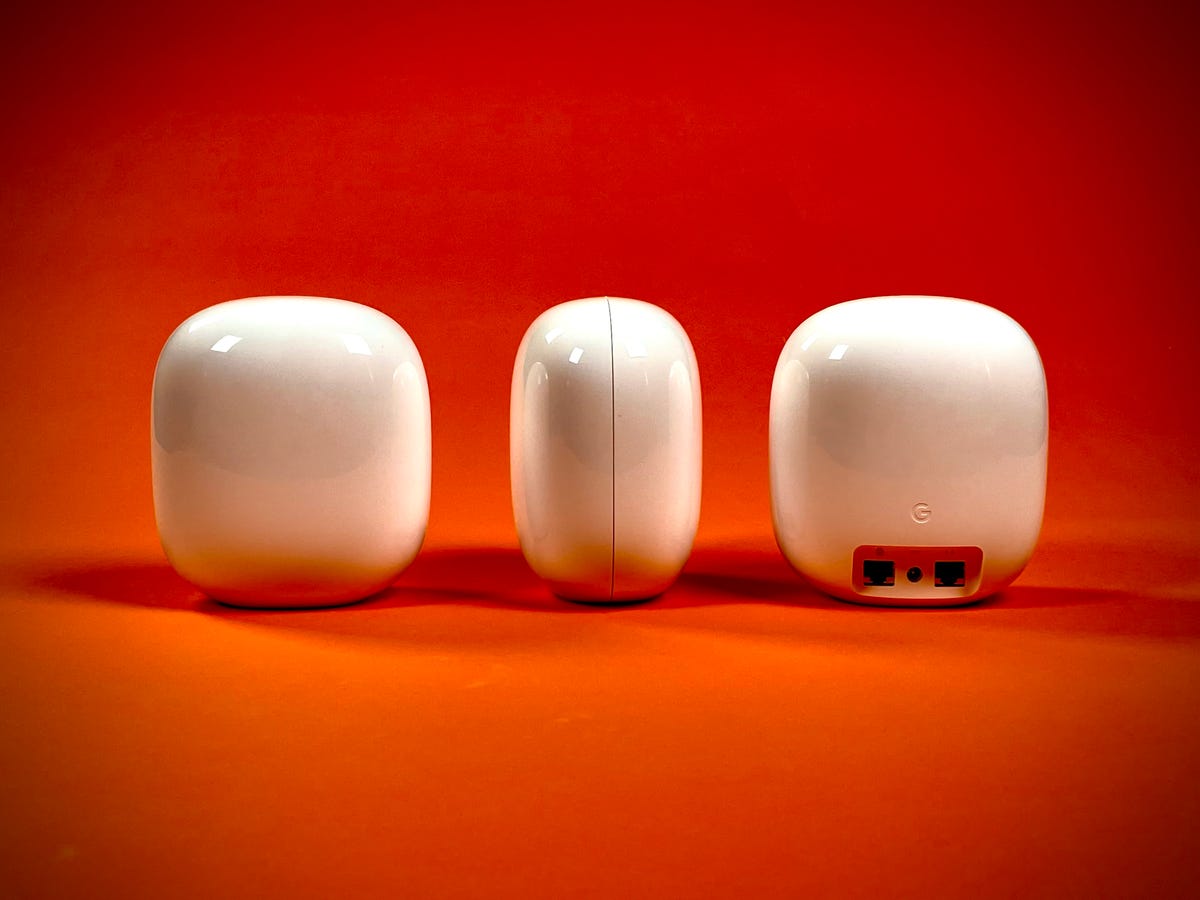 A trio of white-colored Nest Wifi Pro mesh router devices against a fiery orange background. The first is front-facing, the second shows off the profile view from the side, the third has its back to us, revealing a power jack and two ethernet jacks.