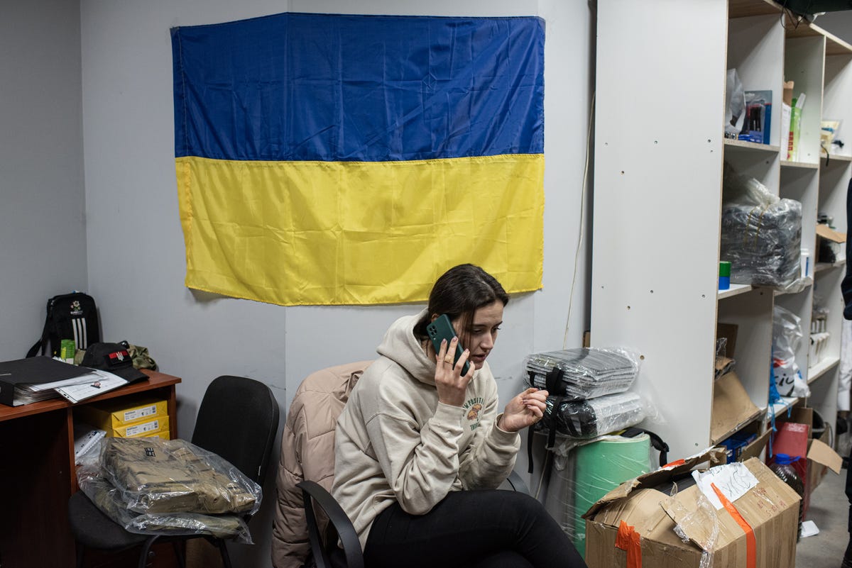 A woman uses a cellphone with a Ukrainian flag hanging on a wall behind her.