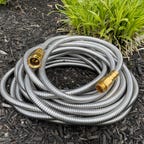 The all-metal Rosy Earth Garden Hose sits in a coil next to some small shrubs.