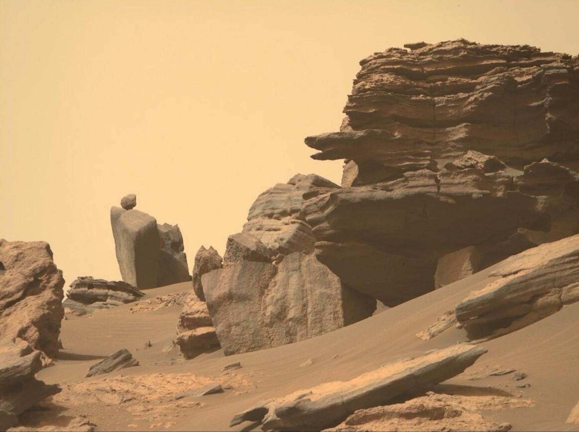A Mars landscape shows a layered cliff face with a nearby small round rock appearing to be impressively balanced atop a larger rock.