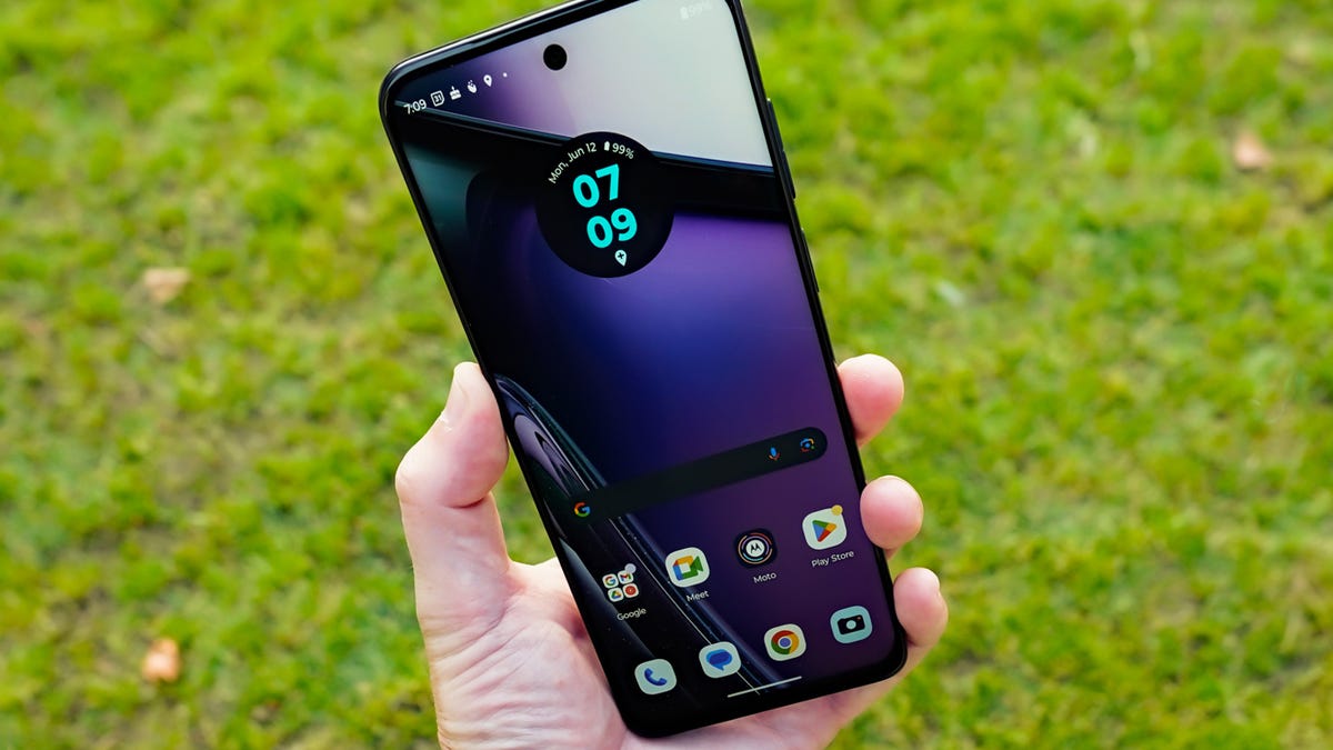 A phone is held up in front of a green grass background, showing the bright display and home Android screen.