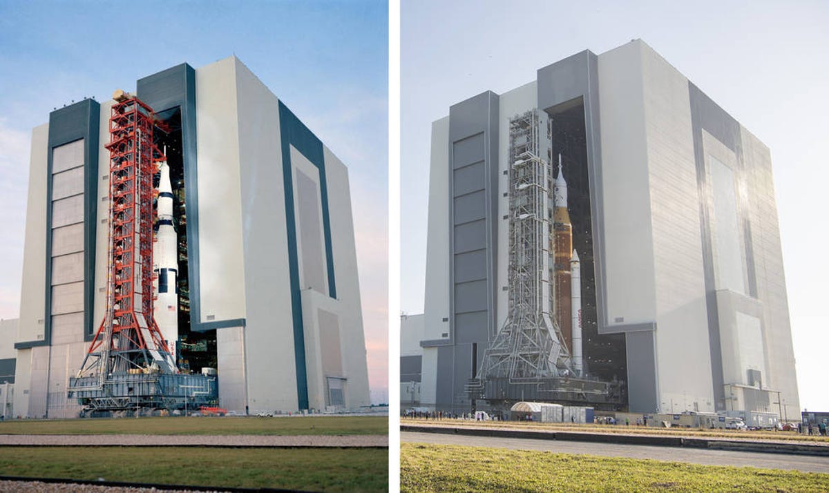 Two massive rockets, Saturn V on the left and SLS on the right, pose in front of the open door of the giant, tall building where they were stacked.