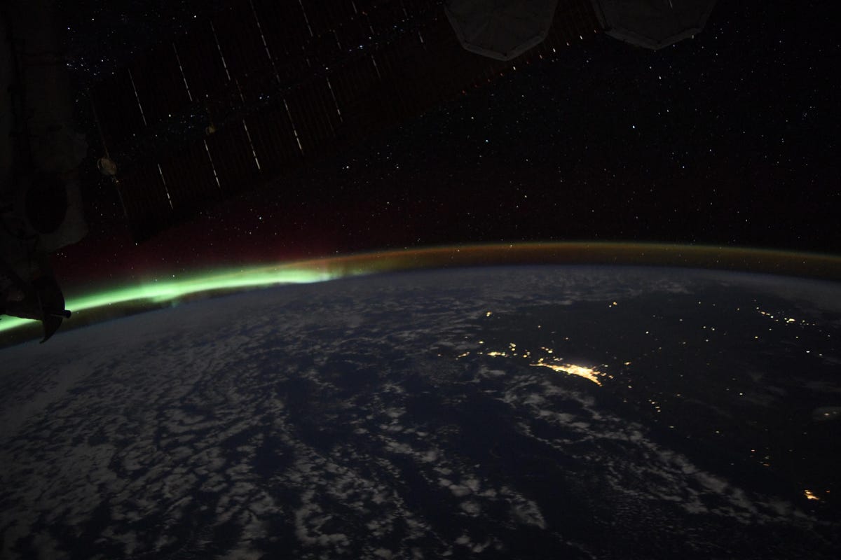 Clouds and city lights are visible on Earth as a glowing green layer of aurora appears in the distance against the darkness of space. A series of light streaks appear in the dark.