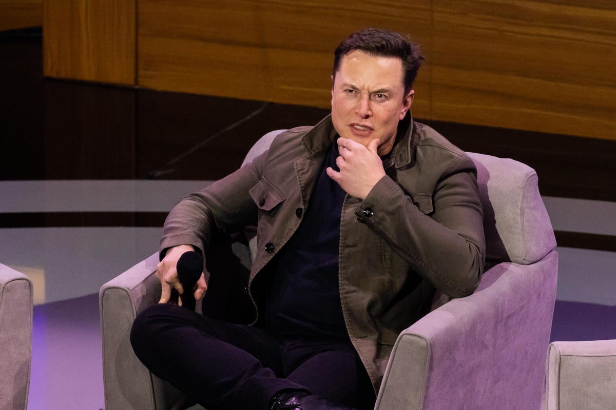 Elon Musk sitting in a purple armchair with his hand on his chin