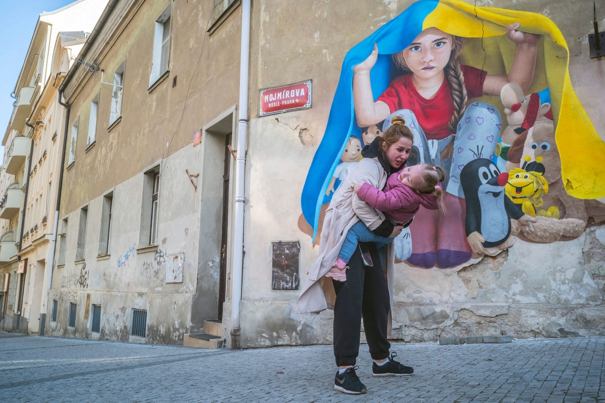 In Prague, a mother holds her child in front of a mural showing a little girl surrounded by toys hiding under a Ukrainian flag. 