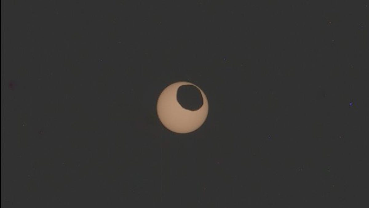 A googly eye-looking view of the sun from Mars with an uneven chunk of Phobos the moon taken out in black.