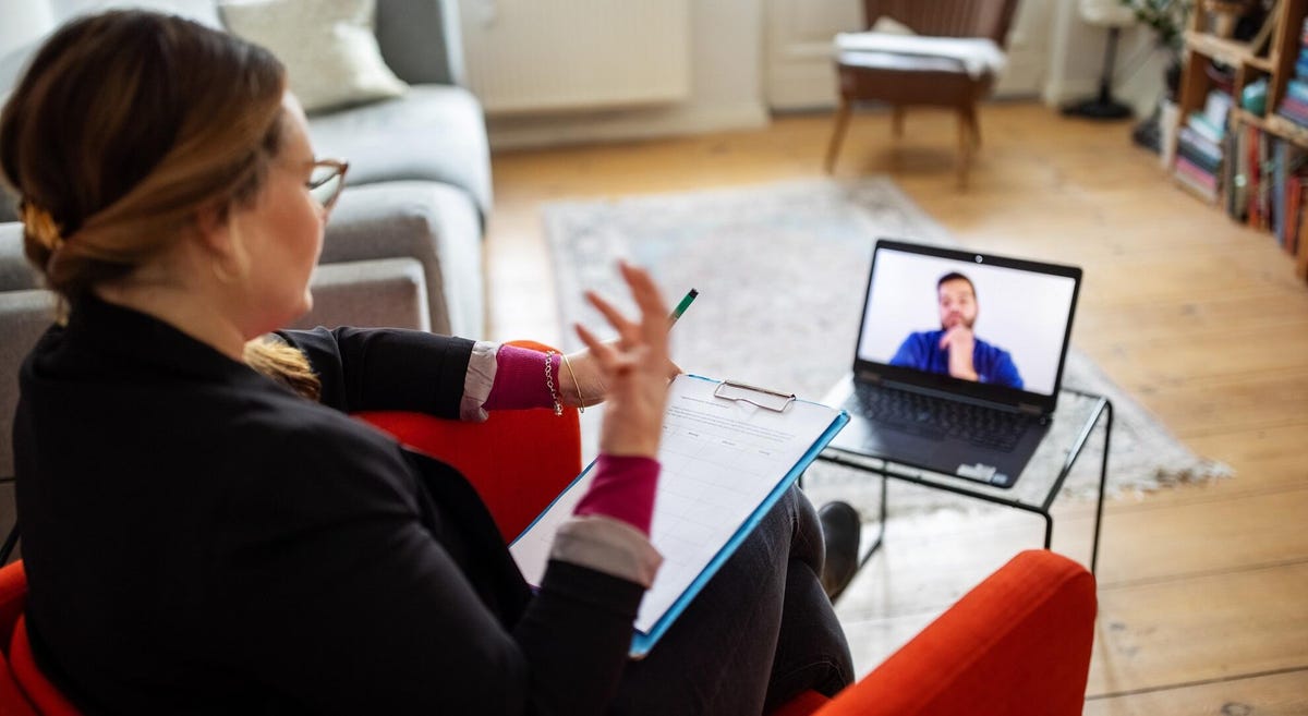 A woman in a red chair holds a clipboard and gestures to the man she's video chatting with on her laptop screen.