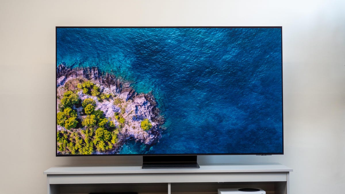 The Samsung S95D OLED TV on a gray TV cabinet in front of a white wall with an image of the ocean and an island on its matte screen.