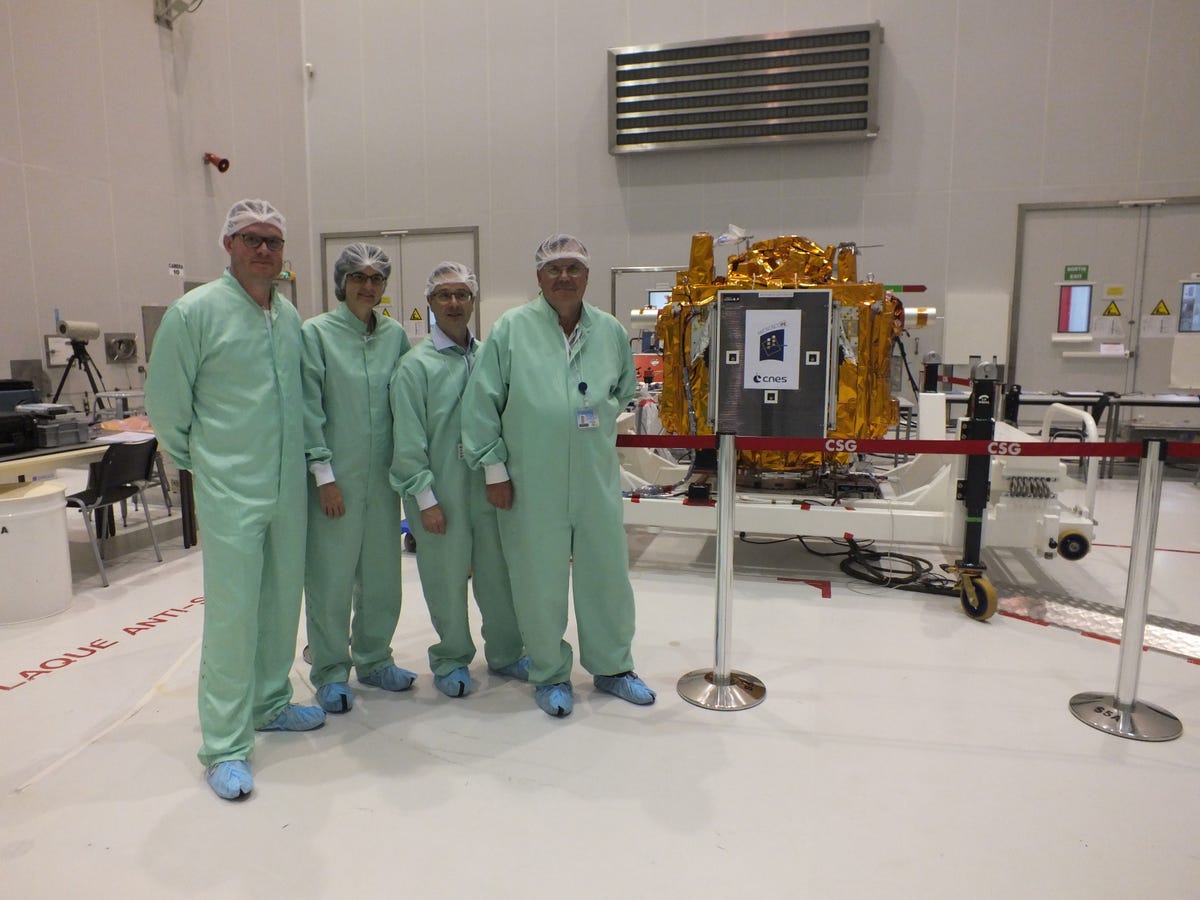Four scientists, in mint green outfits and hairnets, stand next to an oven-sized device wrapped in gold foil
