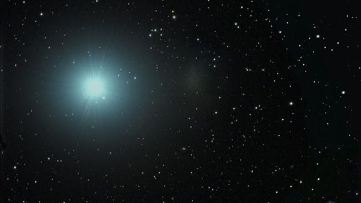 An image of space is dark with speckles of white stars. Toward the left is a soft, hazy green-ish glow.