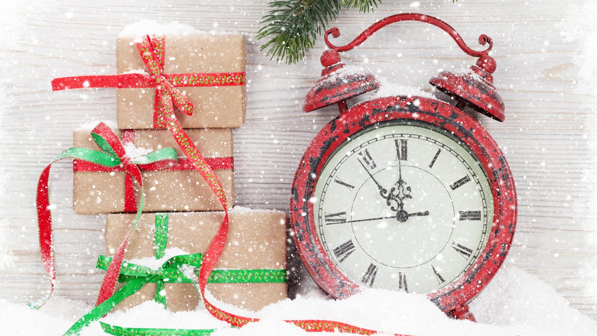 amidst falling snow, an old-fashioned red alarm clock sits next to three stacked holiday packages wrapped in red and green ribbons. the clock shows a time of five minutes before midnight.