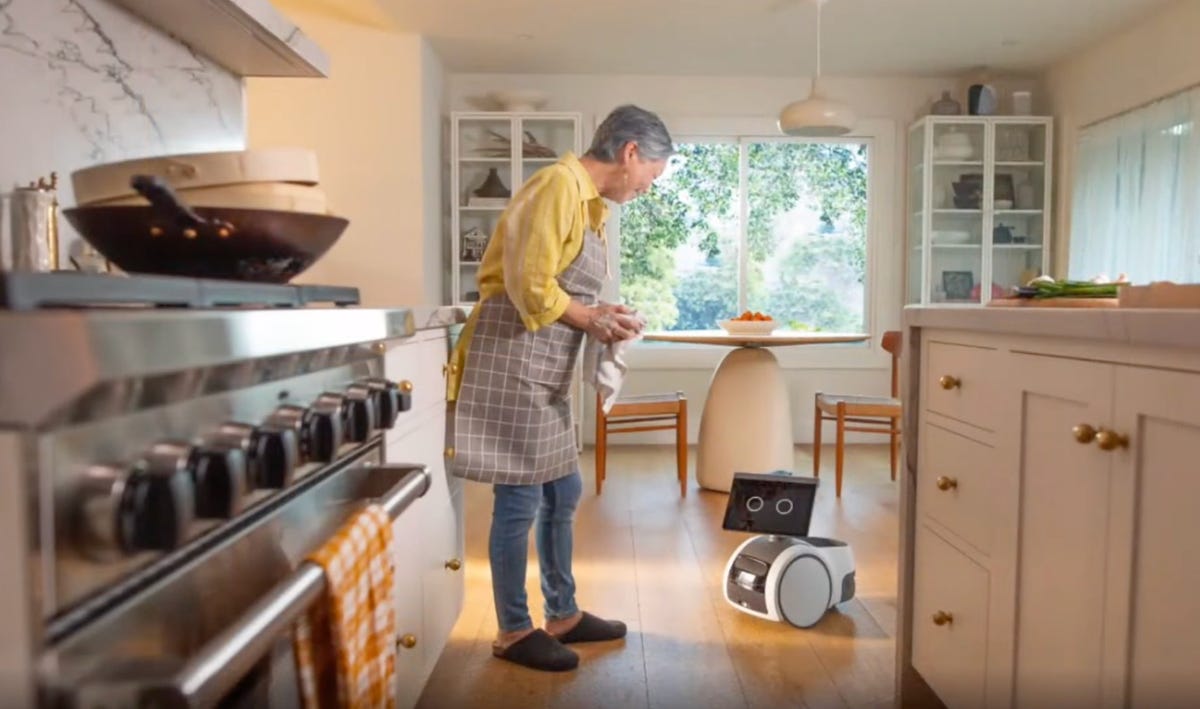 A person wearing an apron in a kitchen leans slightly to address her Astro device