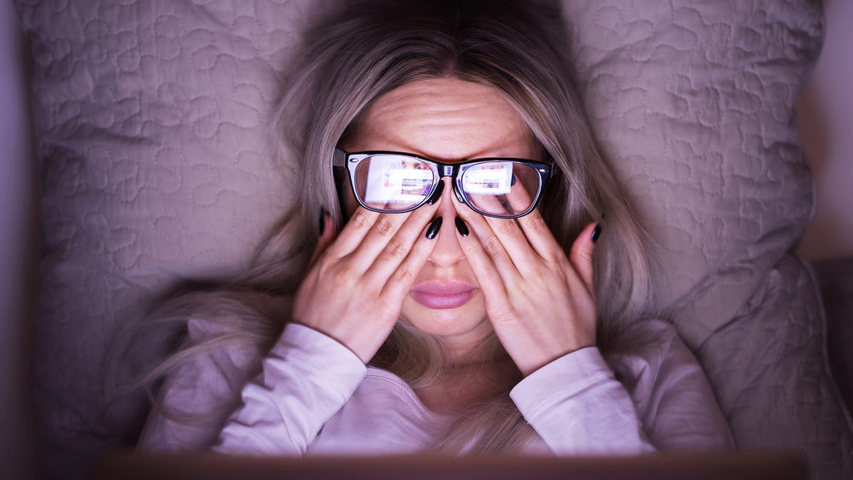A woman with glasses rubs her eyes. The light from a laptop lights her face.
