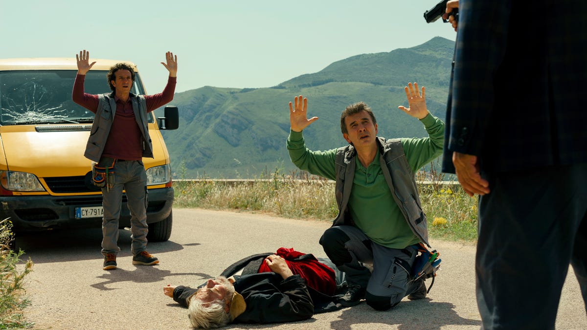 Two men on a road in front of a yellow van, holding their hands up before a man holding a gun. A dead body is lying on the road next to them