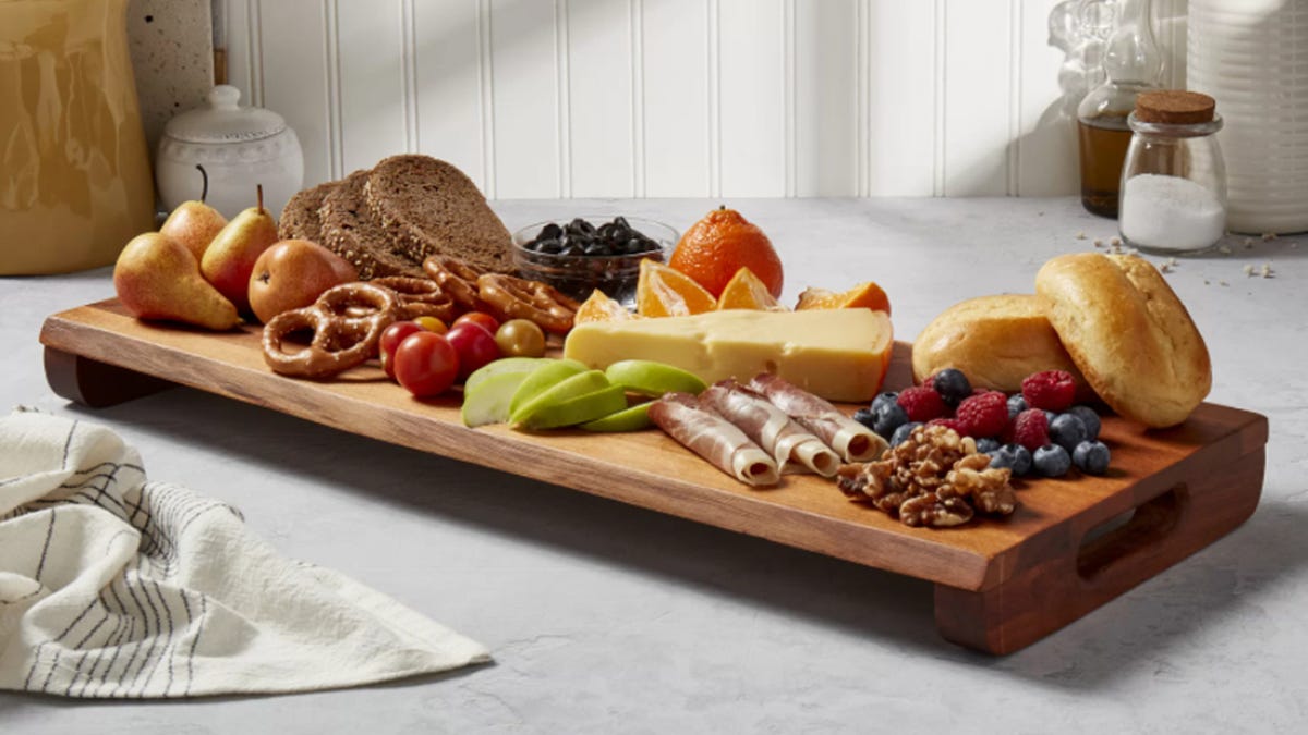 Rich acacia wood cutting board with fruits, meats, breads, nuts and cheeses on a marble counter.