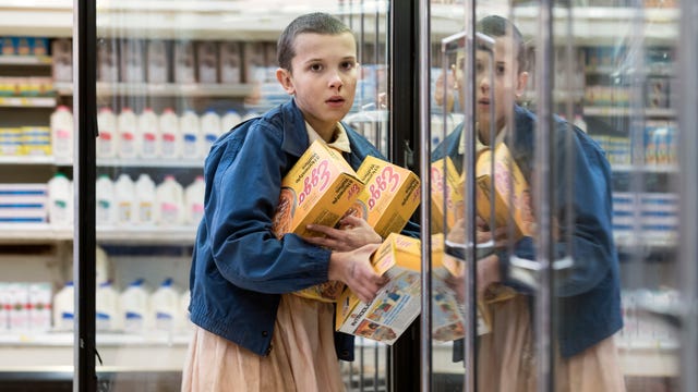 Stranger Things character Eleven holds open a grocery store freezer with her arms full of Eggo Waffle boxes.