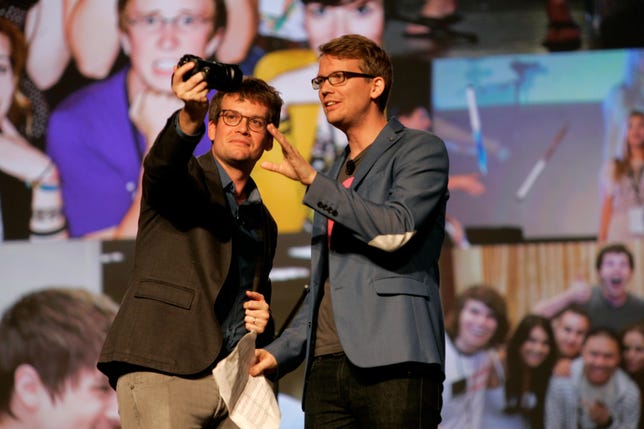 Hank Green and John Green stand on stage, shooting themselves with a video camera