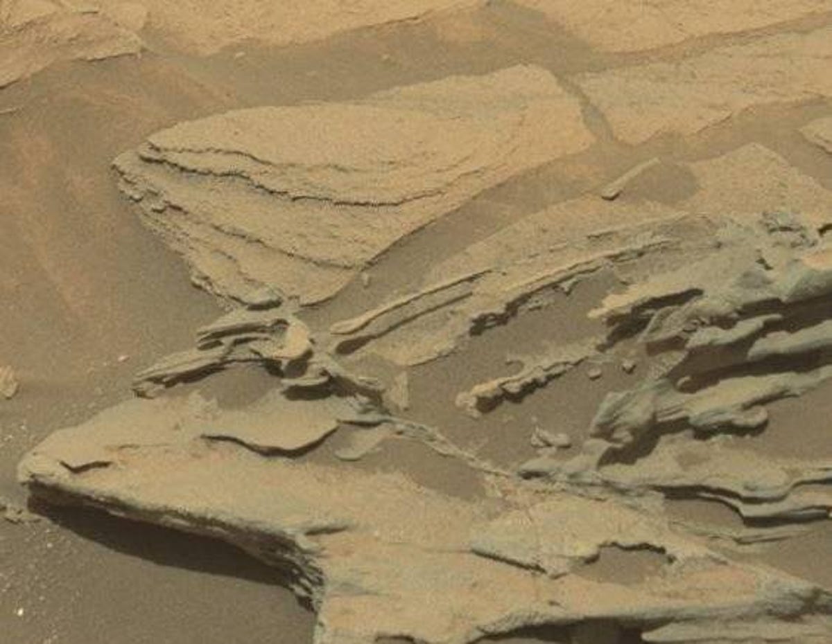 Thin spoon-like formation extends out from beige, sandy Martian rock formation.