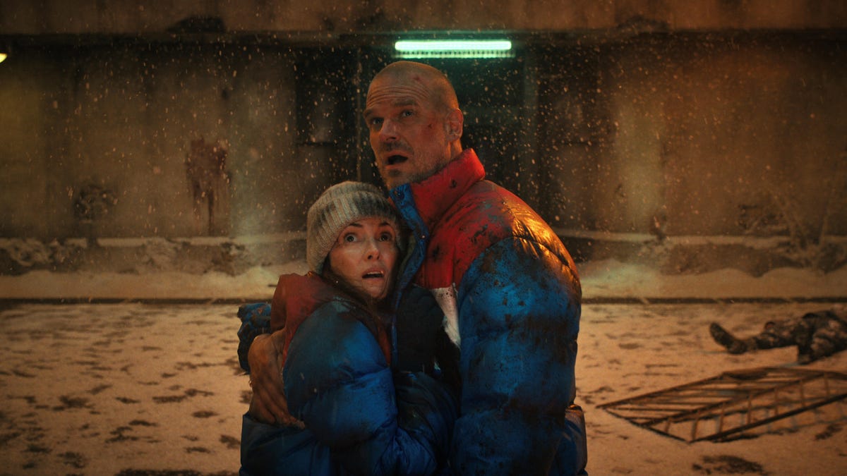 Characters Joyce and Hopper embrace in puffy coats surrounded by bloodied snow.