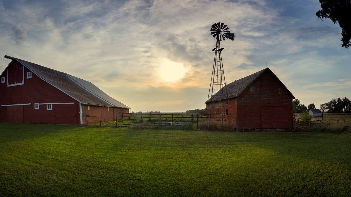 A barn is seen in the foreground with the sun setting behind the clouds in the background.