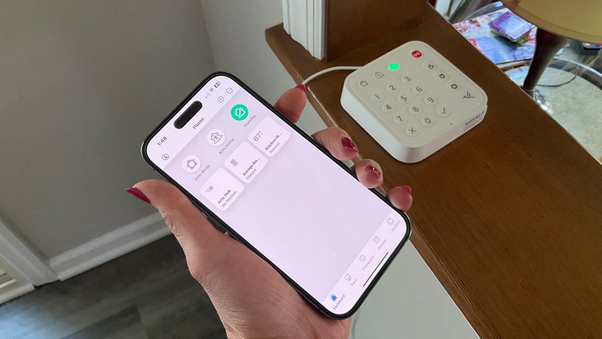 A woman's hand holds a phone in the foreground with the Arlo Secure app's homescreen. In the background, the Arlo Home Security System's keypad hub sits nearby.