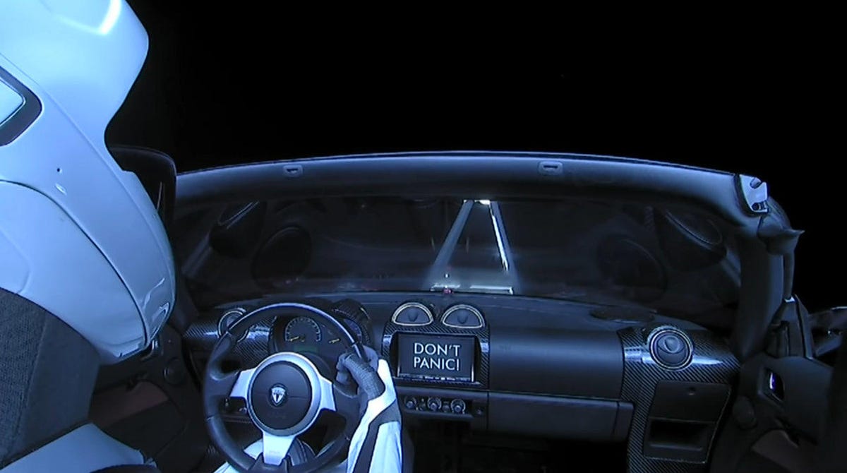 Over-the-shoulder view of helmeted Starman mannequin with Tesla dashboard ahead and darkness beyond.