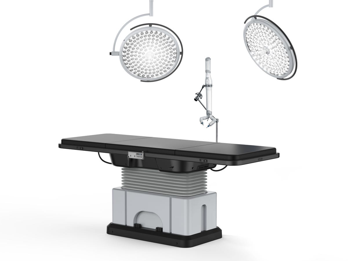 A black patient bed to which MIRA is attached and hovering above.  Above MIRA there are two surgical round luminaires.