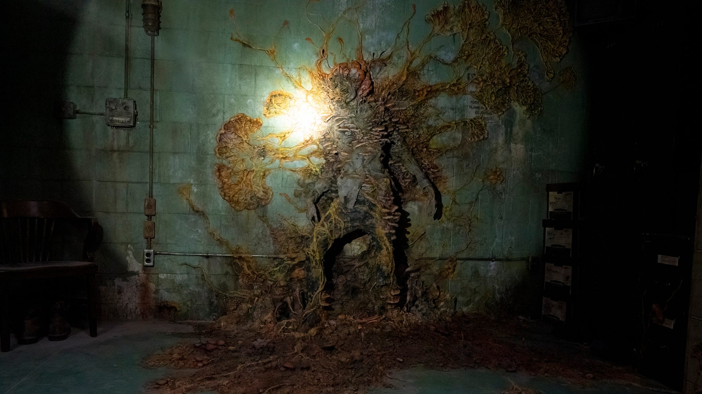 a flashlight shines on a dead body stuck to a wall, spores erupting from it