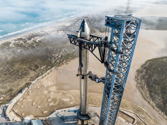 Shiny side of SpaceX's Starship spacecraft angled toward the camera, stacked on a long shiny rocket with launch tower beside it and Texas in the background.