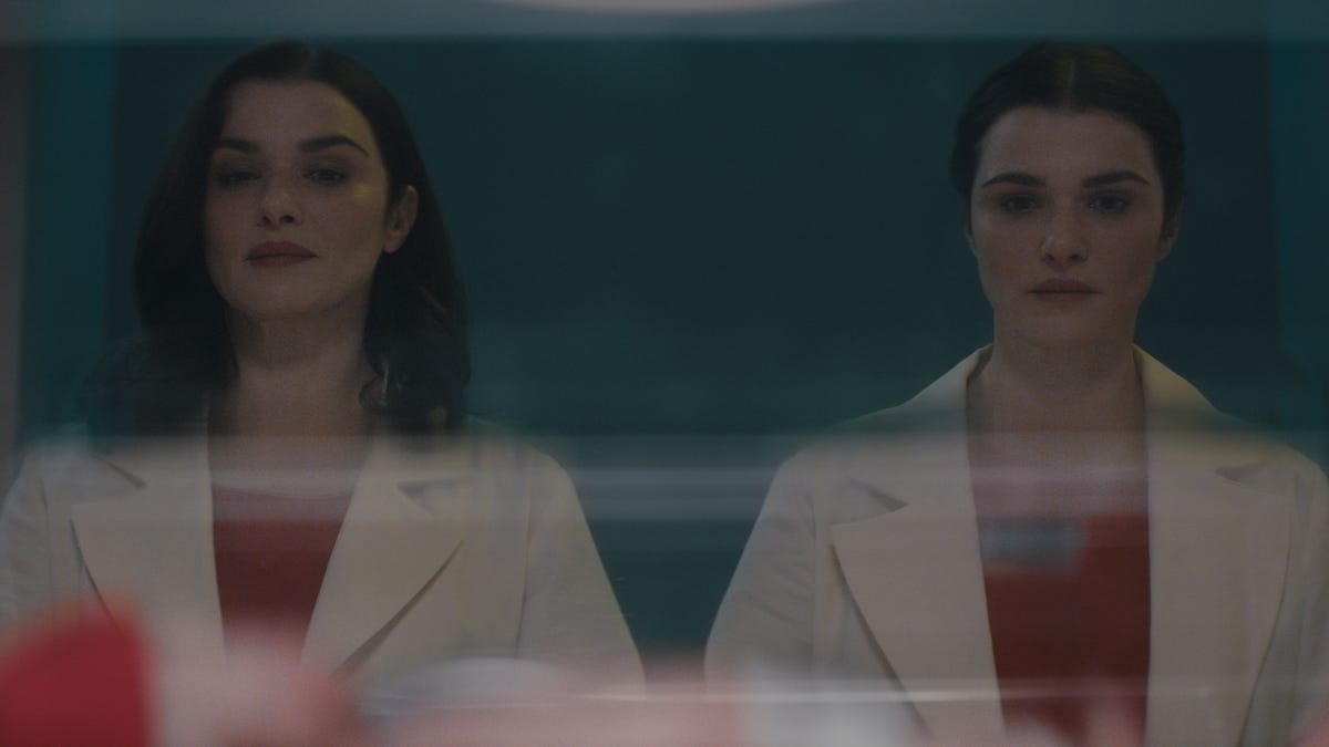 Rachel Weisz in a lab coat and red shirt standing in front of a mirror next to herself with her hair up