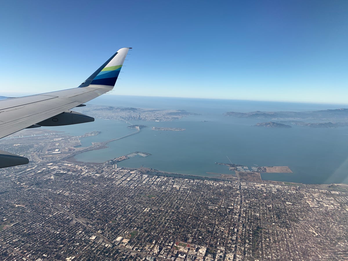 A daytime photo taken by a passenger on a commercial airliner. In the upper left there's the tip of the plane's wing. Down below, the city of Berkeley, California, looks tiny as the San Francisco Bay stretches off into the distance toward the Golden Gate and the open ocean.