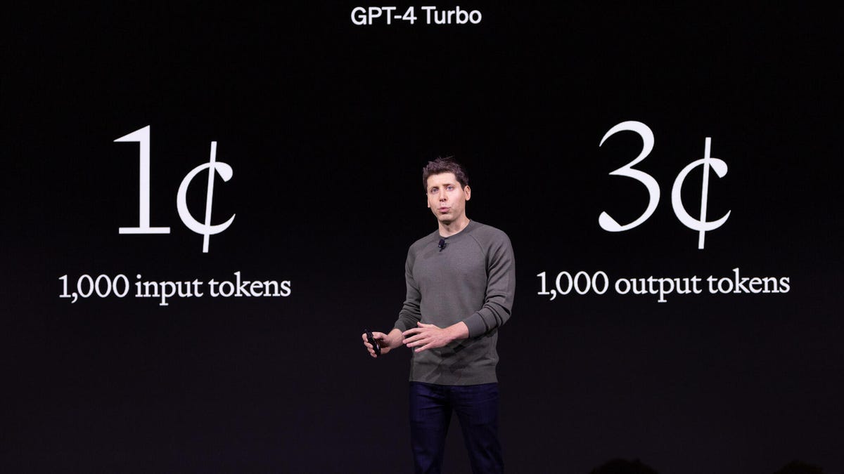 OpenAI CEO Sam Altman speaks at his company's DevDay event in front of a presentation slide showing new lower costs for developers to build tools with the company's GPT AI technology.