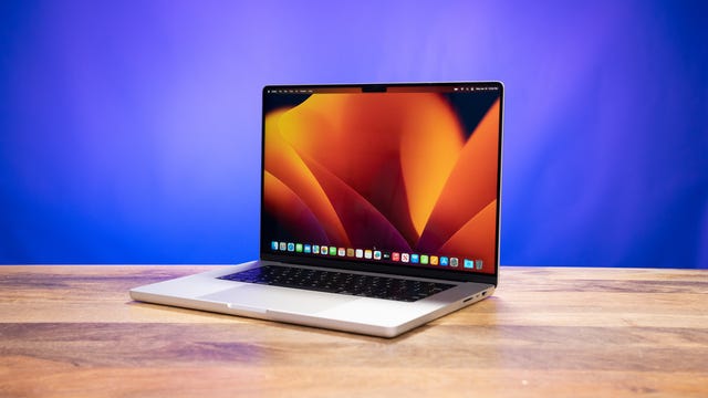 O Apple MacBook Pro 16 sobre uma mesa de madeira, aberto e inclinado para a esquerda, mostrando a área de trabalho em forma de flor de laranjeira contra um fundo azul-púrpura
