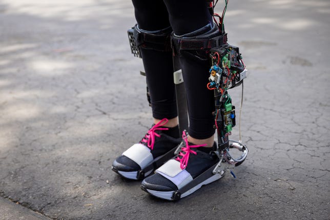 A closeup view of a women's legs, wearing a portable ankle exoskeleton boot (developed by Stanford University's Biomechatronics Lab) strapped to each calf.