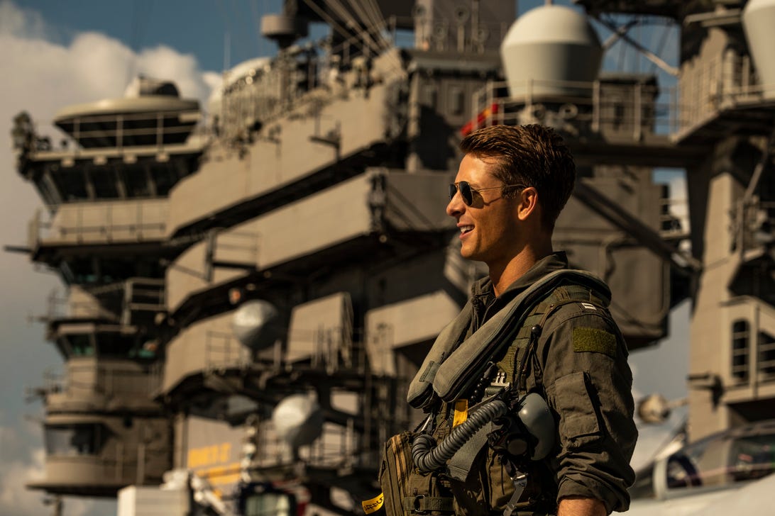 Actor Glen Powell grinning in military flight and shades o the flight deck of a US Navy aircraft carrier.