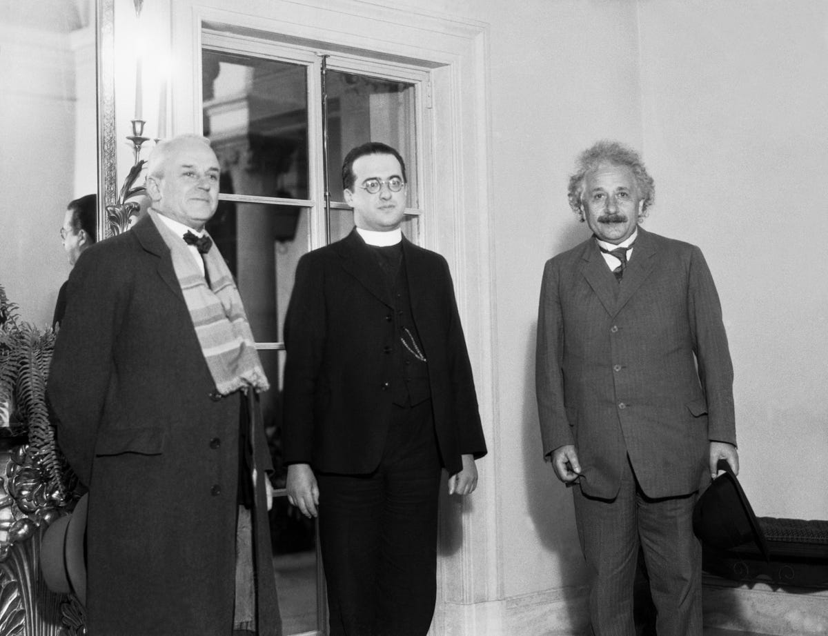 Kairėje yra Millikanas, centre - Lemaître, o dešinėje - Einšteinas.  Trys stovi priešais langą.  Vaizdas yra nespalvotas.