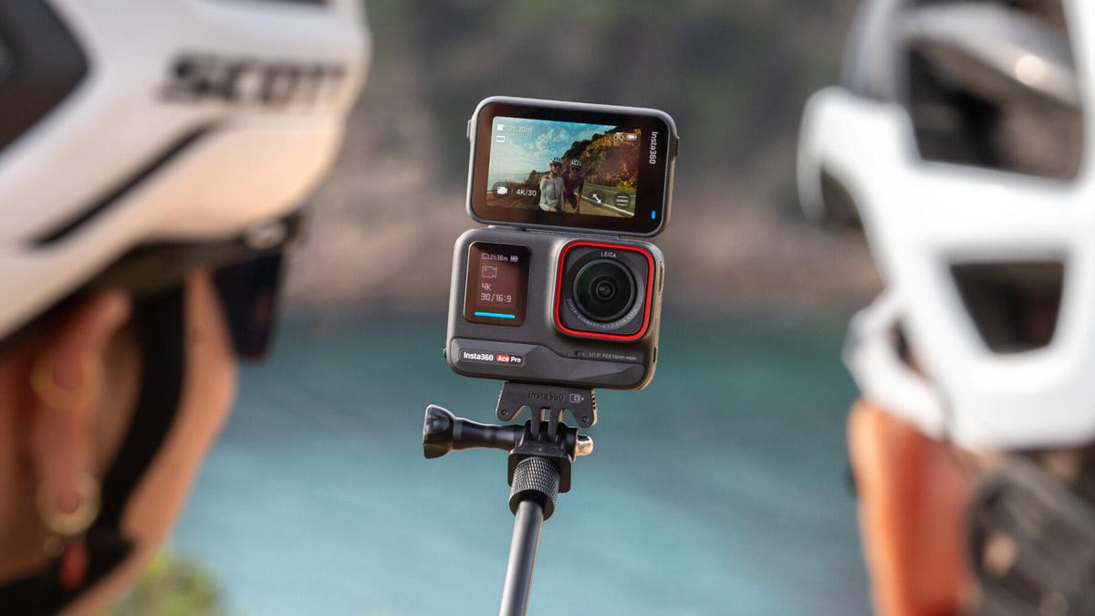 The Insta360 Ace Pro action cam with its rear display flipped up. The camera is on a selfie stick capturing two bicyclists.