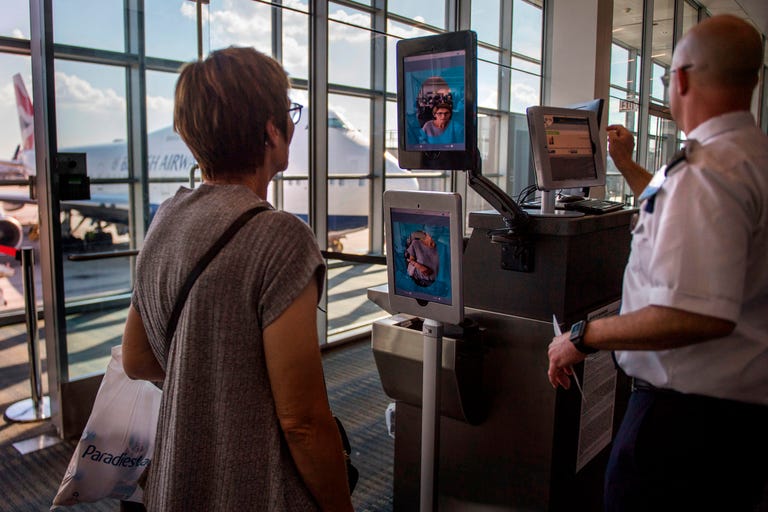 A woman stands in an airport boarding gate in front of a kiosk with two screens showing a photo of her face.