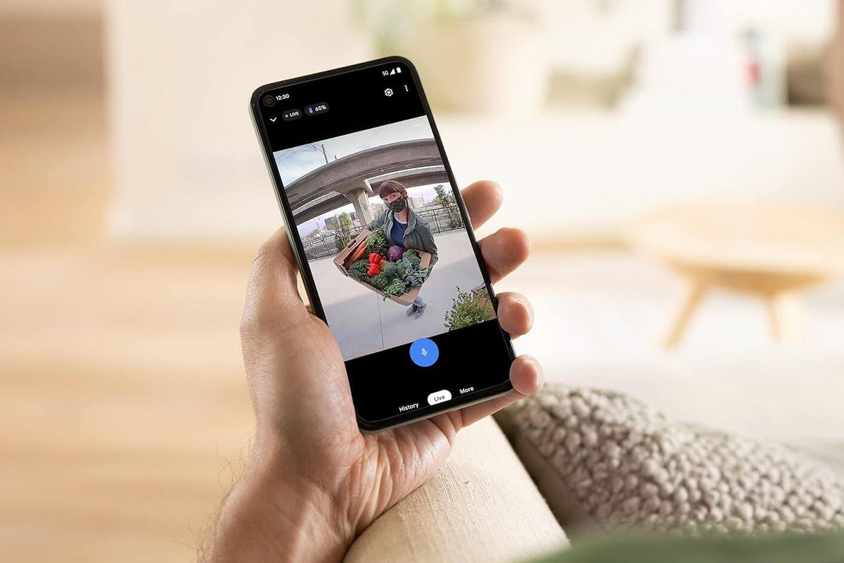 A hand holds a phone to view a Nest Doorbell live view of a delivery person at the front door.