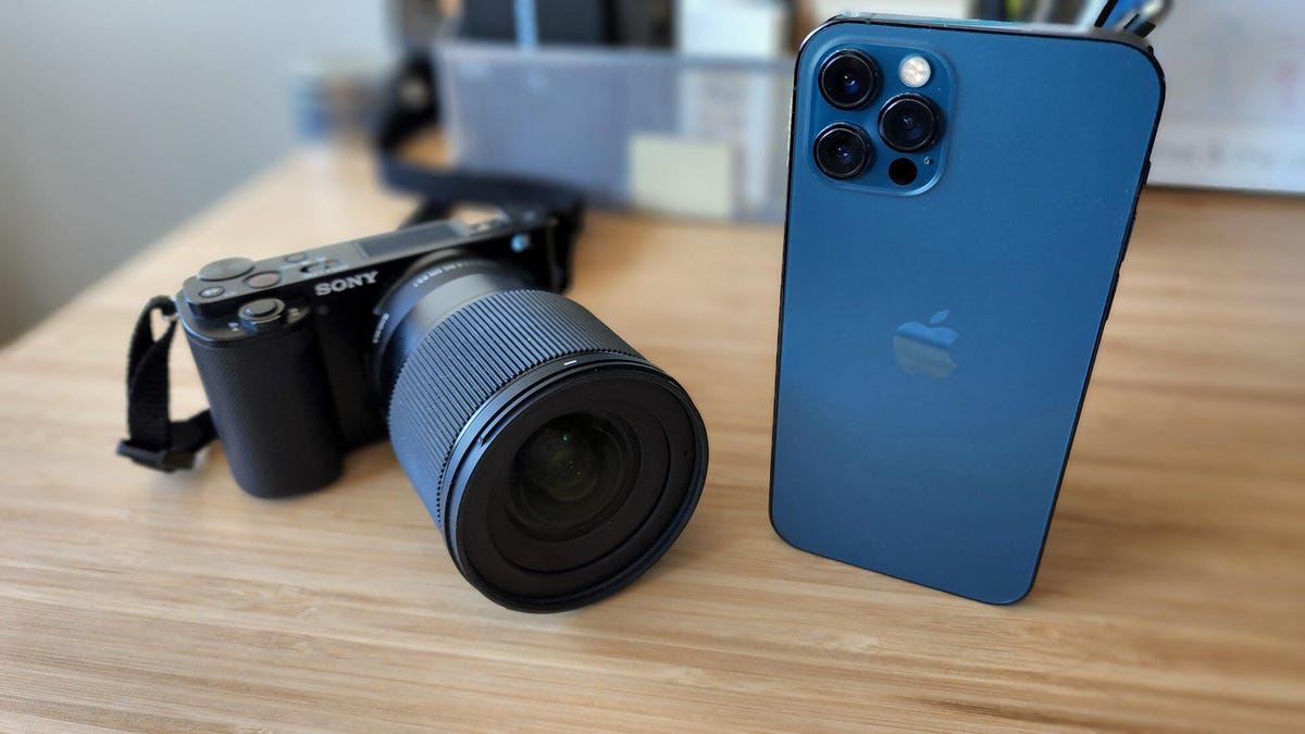 A Sony ZVE-10 mirrorless camera and Sigma 16mm f/1.4 lens sits on a bamboo desk next to a blue iPhone 12 Pro.