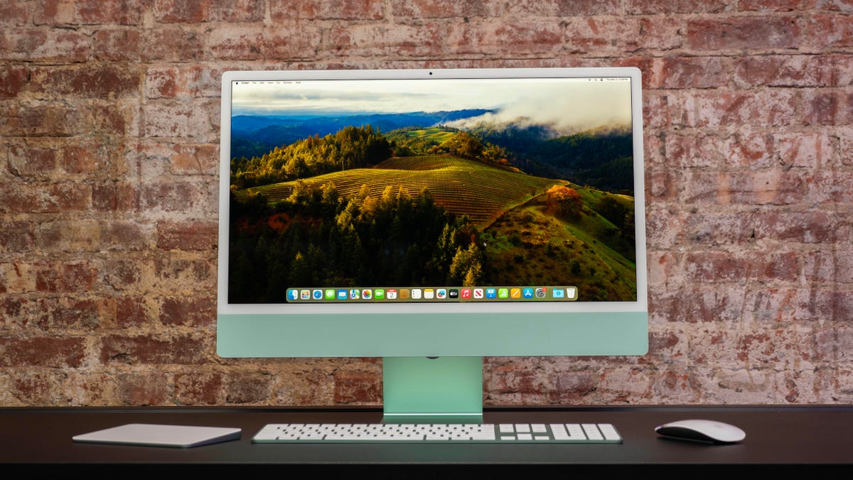 Apple iMac M3 in green with its color-matched accessories in front of it and sitting on a black desk in front of a brick wall.