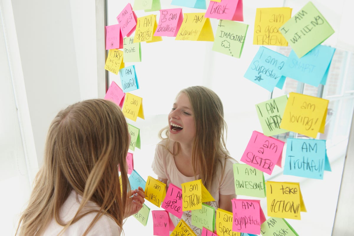 A girl laughs while looking at herself in the mirror and pasting notes with positive affirmations about herself.