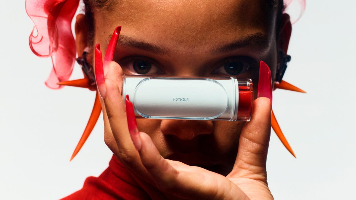 A model looks demurely behind the cylindrical earbuds case she's held between her face and the camera. The case is white, with some translucent covering, and the word 'Nothing' printed in small type. One of the case's ends is mysteriously red.