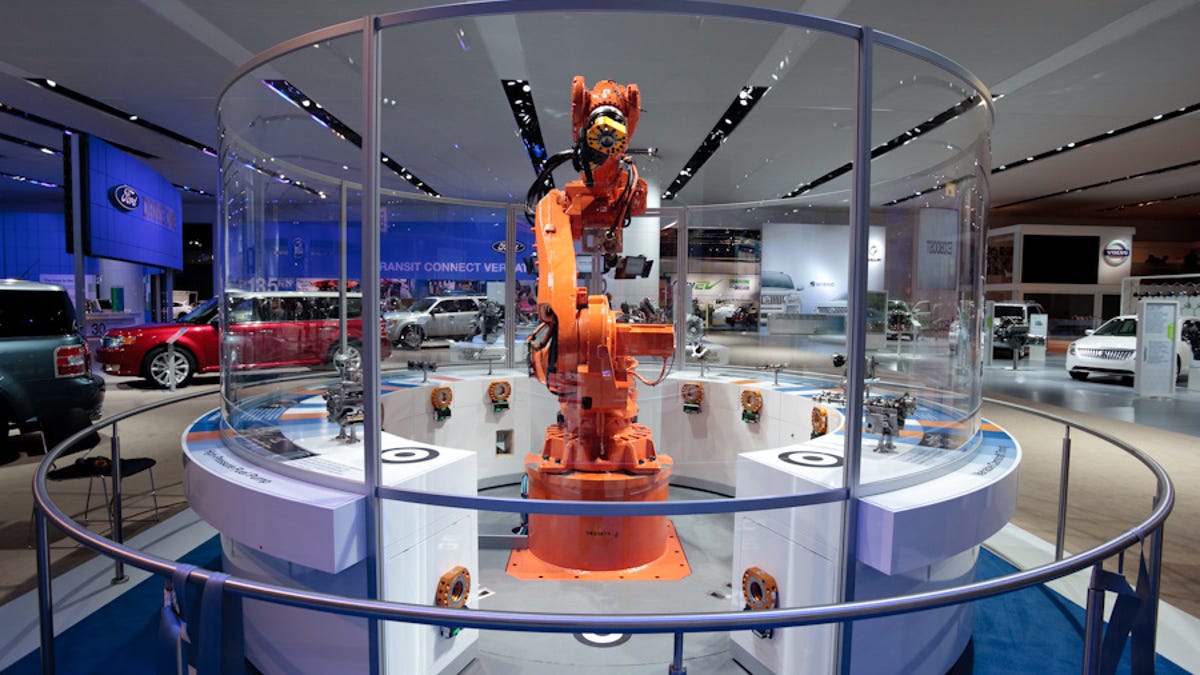 An orange, vaguely human-shaped robot stands mounted in a round glass display case, which is surrounded by a metal railing. Cars are on display in a large show room behind it.