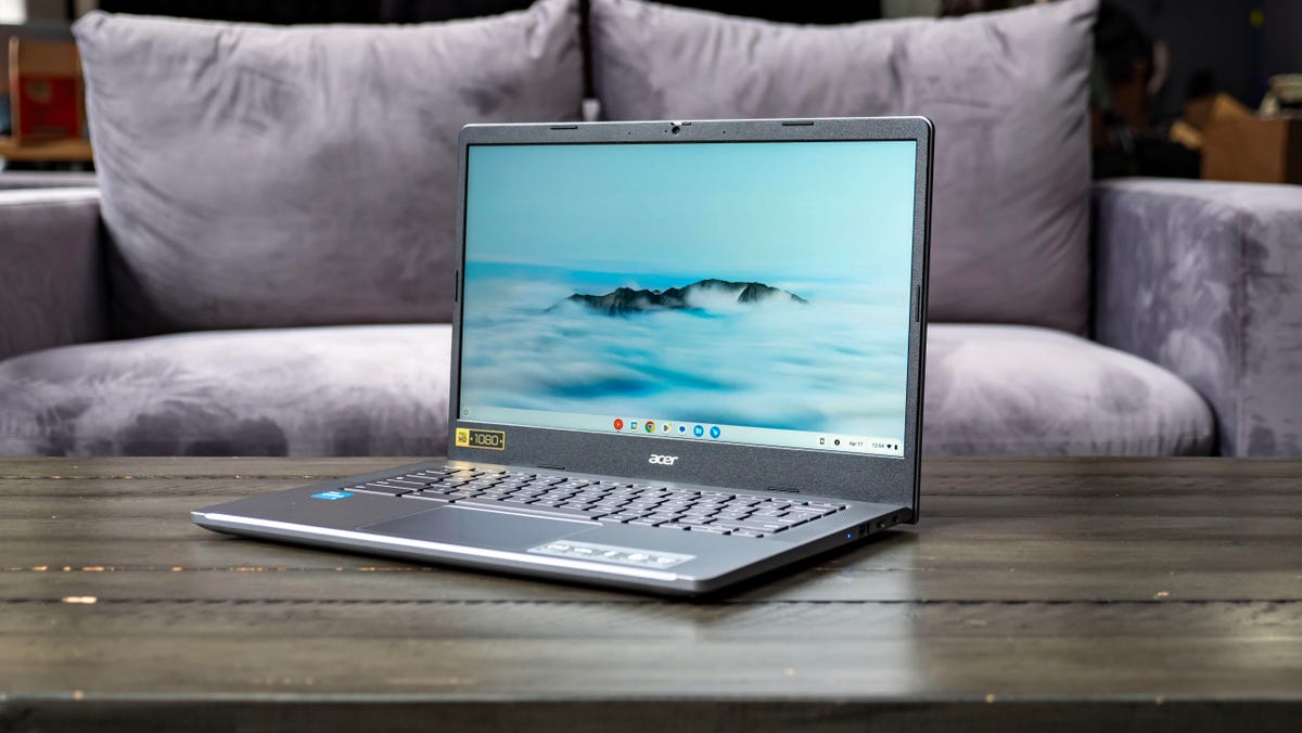 Acer Chromebook Plus 514 aberto e voltado para a esquerda em uma mesa de madeira em frente a um sofá cinza claro.